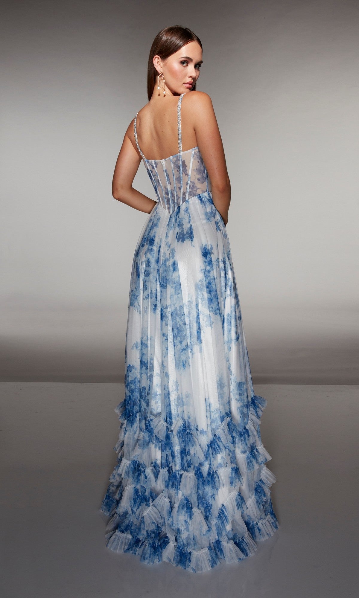 Woman in the Alyce 62202 long prom dress, featuring a sleeveless design, sheer back, blue and white floral patterns, and tiered ruffles, standing with her back to the camera.